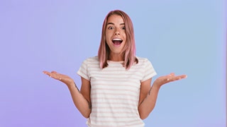 Woman with pink hair and tattooed hands, dressed in striped t-shirt. Looking amazed and smiling, saying wow, posing on purple background. Close up