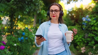 Woman with coffee and smartphone walking through the blooming garden