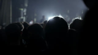 Energetic crowd of people seen from behind, cheering and waving hands at a live music concert or festival. Dynamic stage lights illuminate