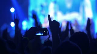 Excited viewers applauding to the singer or band performing on colored illuminated stage. One of the fans taking picture or making video with digital camera