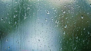 Raindrops fall on a window with an out of focus red light in the background.