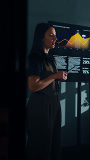 Confident businesswoman presenting analytics data on large screen with colorful line charts during professional meeting in modern dark