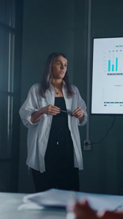 Confident businesswoman in white shirt delivering data-driven presentation with charts and graphs on large screen during modern corporate