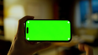 Hands holding modern smartphone with vibrant green screen in cozy dimly lit room, perfect for compositing, mockups, tutorials, or digital