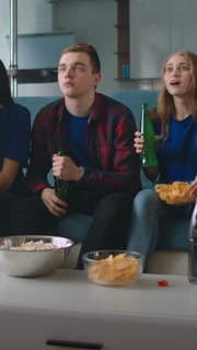 Friends watching an exciting sports game at home, cheering together on a cozy couch with snacks and drinks, celebrating an intense moment in