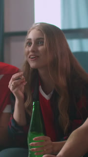 Young woman in casual sportswear cheering and laughing while watching a live match at home, enjoying snacks and drinks in a relaxed social