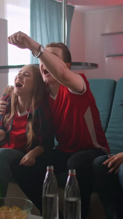 Friends in red sports jerseys sitting on a couch with drinks and snacks, intensely watching a game together and celebrating moments in a