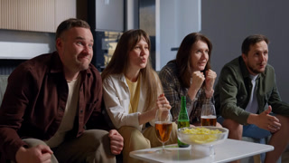 Group of four friends sitting closely on a couch in a cozy living room, enjoying drinks and snacks while engaging in a lively conversation