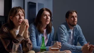 Three adults sitting closely together at a table indoors, watching something with focused expressions that turn into smiles and laughter