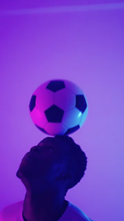 Silhouette of a football player in a white shirt skillfully balancing a soccer ball on his head under purple and blue lighting in a