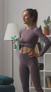 A woman in fitted athletic wear staying hydrated while taking a break from her home workout routine in a cozy living room environment