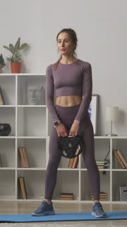 A young woman performing a strength training workout at home in her living room using a weight plate, wearing coordinated athletic wear and