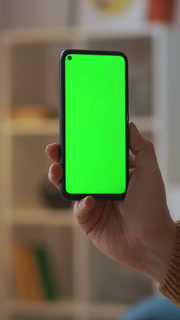 Hand holding modern smartphone with green screen in indoor setting, showcasing blank display for digital content or app presentation in
