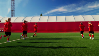 Womens soccer team warming up on modern training pitch, athletes in red uniforms practicing running drills together under clear blue sky