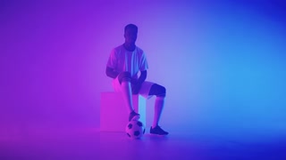 black football forward of halfback is sitting on cube in studio, posing with ball