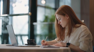 redhead woman student is learning remotely, doing homework in exercise book, sitting at table with laptop