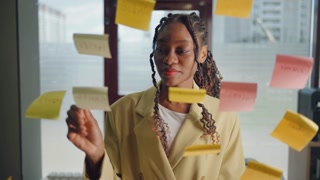 Smalling African american business woman using sticky notes brainstorming problem solving strategy on glass whiteboard, leader woman showing solution for project deadline in office.