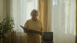 Medium full shot of older woman with white hair flipping through vintage photo albumwhile walking beside window in retro living room