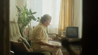 Side view of senior woman with grey hair looking through photo album reminiscing bittersweet memories of youth then leaning back on couch in retro living room