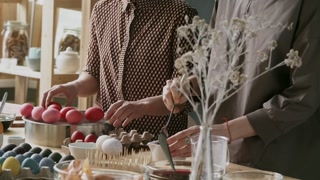 Tilt down of Asian woman putting colored eggs on tray as her female friend with short hair coloring Easter egg in red dye