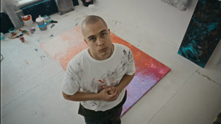 Portrait of Caucasian young adult man standing in art studio next to abstract painting on floor, looking up at camera. High angle shot ideal for creative industry promos or art school marketing