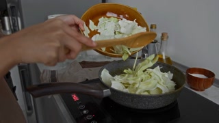 Close-up shot of hands of unknown woman putting cabbage leaves in frying pan using wooden utensil when cooking in kitchen