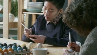 Medium shot of Asian woman and her female friend drawing with pens on colored Easter eggs