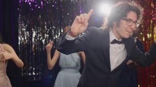 Cheerful teenage guy in tuxedo and eyeglasses looking at  camera while dancing against group of intercultural friends enjoying prom party in night club