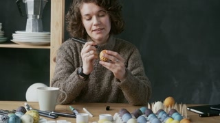 Medium shot of happy young woman with short curly hair using marker and drawing on colored Easter egg
