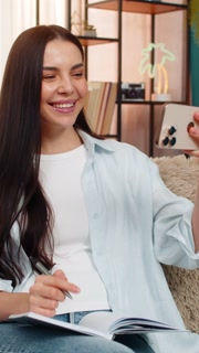 Young woman on sofa wearing headphones watching video lesson on mobile phone taking study notes