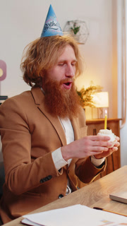 Happy business office man wears festive birthday hat hold cupcake makes wish blowing burning candle