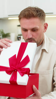 Vertical view of excited Young Caucasian man lifts lid of gift box and smiles with joy at home