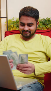 Smiling happy man counting money cash and use laptop pc calculate domestic income earnings at home