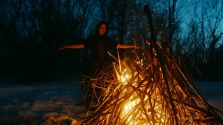 Witch performing witchcraft at bonfire. Mysterious female conjuring in forest during wintery night. She is doing rituals on snow covered