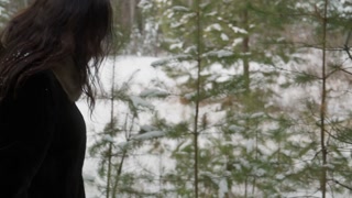 Woman in black fur coat crouching near small snow-covered pine tree in forest. Observing nature, gentle interaction with snowy branches