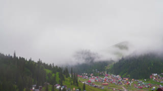 A wide, static shot shows a remote village with colorful houses nestled in a green mountain valley. Thick white fog and low clouds drift