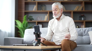 Confident elderly senior gray haired male private tutor giving video call lesson or training using smartphone sitting on sofa in living room at home. Blogger is streaming. Teacher has an online course