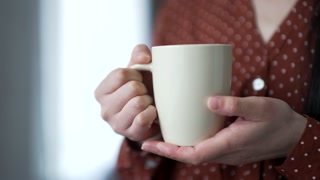 Close up shot of a woman's hands holding a cup of coffee indoors at home. Young Asian woman enjoying a hot drink in the morning in a cozy living room or kitchen. Girl in shirt has a white coffee mug