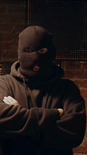 A man in anonymous mask stands against a brick wall. Bad guy