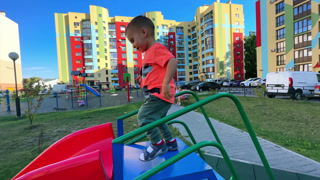 Caucasian toddler climbs on the slide. Baby boy slides down quickly. Bright residential houses at backdrop. Caucasian toddler climbs on the slide. Baby boy slides down quickly.
