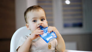 Lovely Caucasian toddler holds a pack of fruit puree in his hands. Baby tries to get some food from the pack with his finger. Close up. Blurred backdrop.