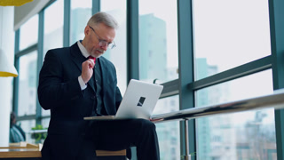 Senior businessman in suit working near office window. Elderly elegant man is typing on a laptop while sitting near the window. Business concept.  Senior businessman in suit working near office window.