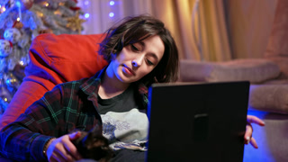 Young Woman with Cat Using Laptop At Home Charming close-up of a smiling young woman using a laptop in a cozy, festively decorated home. She has short brown hair and wears a flannel shirt. She is sitting on a red chair with a black cat sitting next