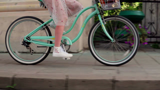 Woman riding a vintage cruiser bicycle with a basket on a sunny day along a city street sidewalk