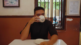 A man drinking coffee while talking to the camera in a cafe, creating a point of view conversation as if on a casual meeting or date. Warm and friendly atmosphere with direct eye contact and social interaction.