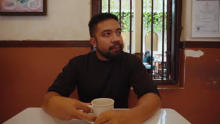 A man drinking coffee while talking to the camera in a cafe, creating a point of view conversation as if on a casual meeting or date. Warm and friendly atmosphere with direct eye contact and social interaction.
