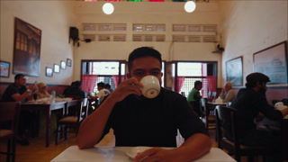 A man drinking coffee while talking to the camera in a cafe, creating a point of view conversation as if on a casual meeting or date. Warm and friendly atmosphere with direct eye contact and social interaction.