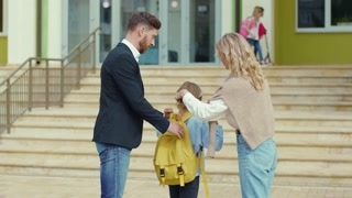 Affectionate young parents with lovely daughter entering the school. Adorable little elementary girl saying goodbye to parents wearing