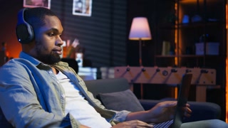 African american freelancer listening to a podcast being focused on online work, using headphones while seated on the couch in his living