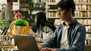 Young man typing on laptop in library study space and learning online, classmates work nearby with books and shelves in soft focus around